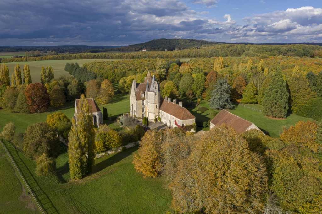 Photographie du manoir de la Vove à Corbon réalisée avec un drone. Dans la vallée de l’Huisne, entre Mauves et Boissy-Maugis, se dresse la sil- houette imposante de la Vove. Le manoir n’est pas bâti sur une hauteur, ni même, comme souvent, à flanc de coteau. Il est en terrain plat, presque en fond de vallée, dans une région autrefois couverte de forêts. La Vove est l’un des plus importants manoirs du Perche. Le site est connu depuis le XIIe siècle mais le manoir, tel qu’il apparaît aujourd’hui avec sa chapelle, mêle le gothique de la fin du XVe siècle à la recherche d’un certain confort lors d’une importante campagne de travaux réalisée au XVIIe siècle. C’est la raison notamment de la présence de ces fenêtres agrandies dans le but de laisser entrer davantage de lumière. Les seigneurs de la Vove ont longtemps compté parmi les plus puissants du Perche. Ce n’est qu’au moment de la Révolution, lorsque le manoir fut vendu comme bien national, qu’il devint une ferme. Il ne cessa alors de se dégrader jusqu’en 1968 ou un passionné en fit l’acquisition, le sauva d’une ruine très probable, et entama une restauration exemplaire. Photographie extraite du projet photographique "les manoirs du Perche"